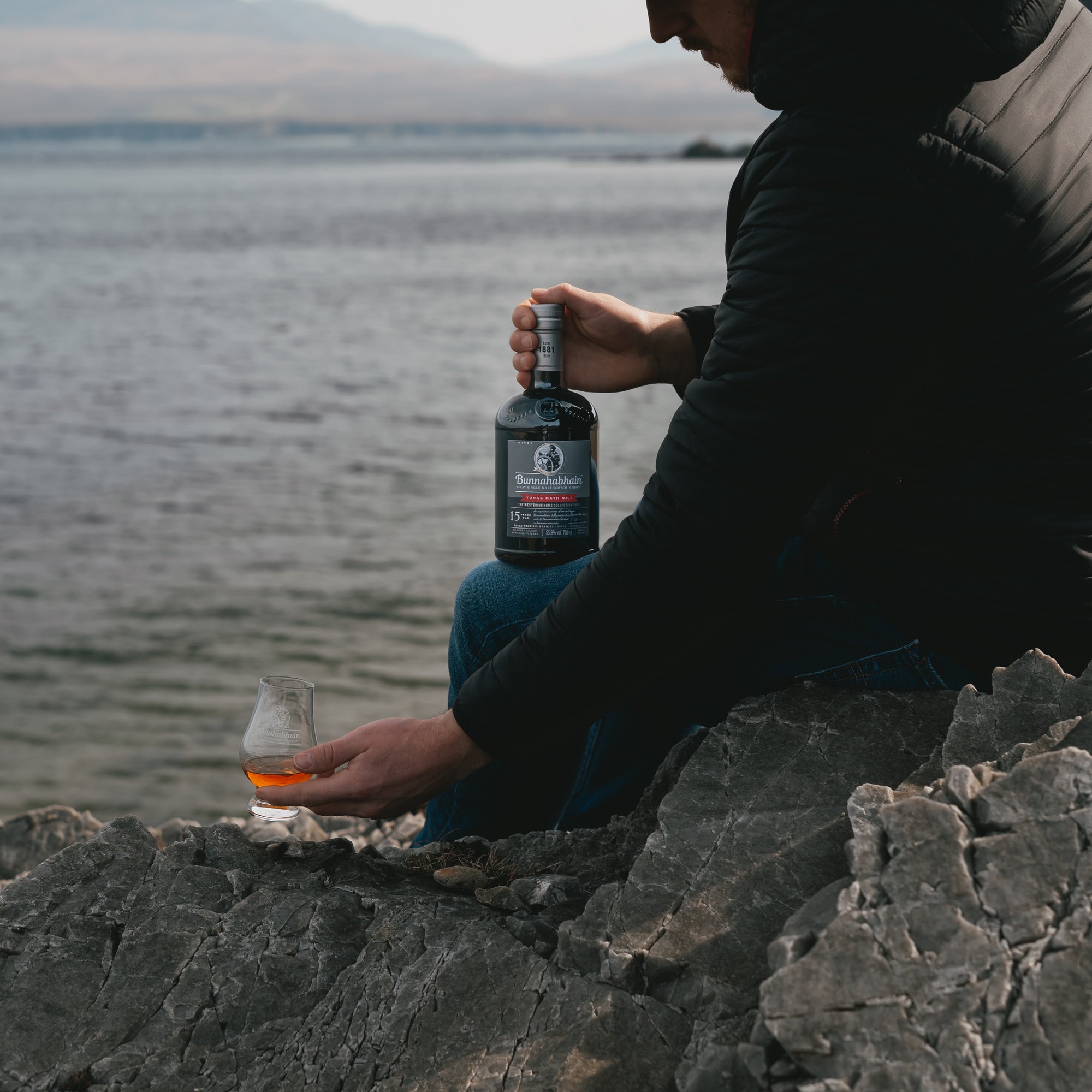 A man sitting on rocks with a bottle of Bunnahabhain Turas Math No.1 and picking up a dram.