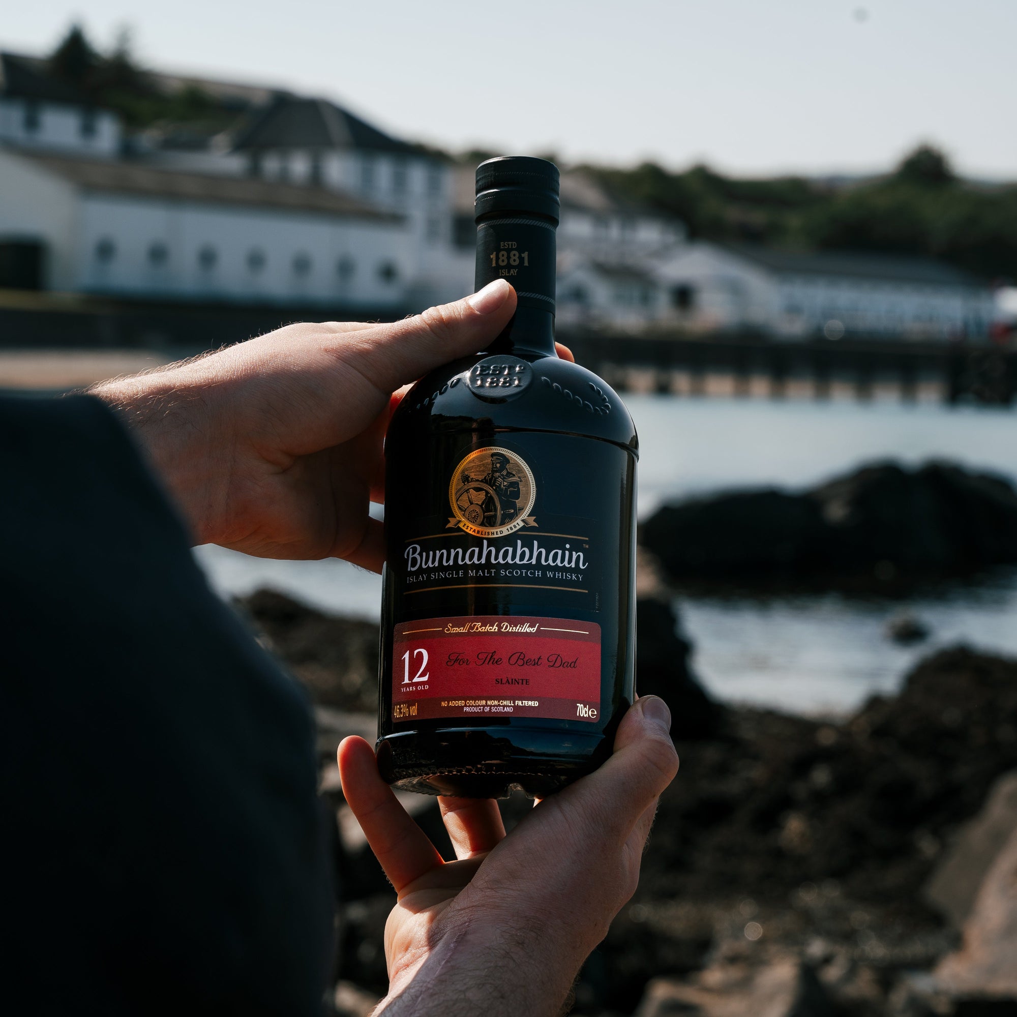 Person holding a bottle of Bunnahabhain whisky by a coastal scene