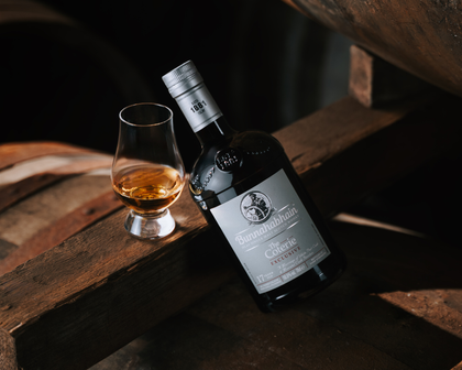 A bottle of Bunnahahain 17 Year Old White Port and Moscatel Cask Finish is angled against a cask in a warehouse setting. There is a Glencairn glass next to the bottle with whisky in it.