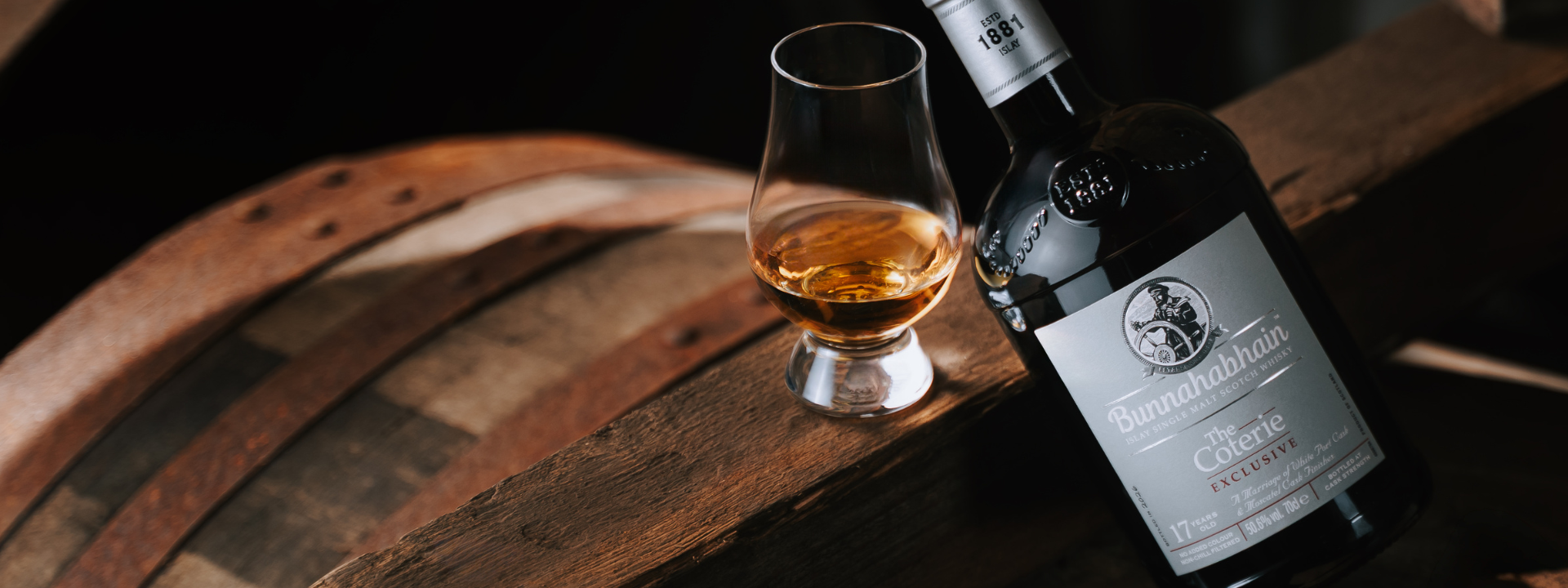 A bottle of Bunnahahain 17 Year Old White Port and Moscatel Cask Finish is angled against a cask in a warehouse setting. There is a Glencairn glass next to the bottle with whisky in it.